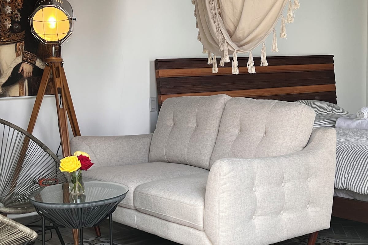 Una habitación de hotel moderna con un sofá gris, una mesa redonda de vidrio con un jarrón de flores, una lámpara de pie de madera y una cama con ropa de cama a rayas.