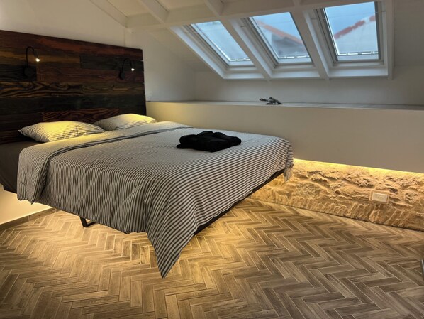 A bedroom with a herringbone floor, a bed with striped bedding, a headboard with a wooden panel, and skylights above the bed.