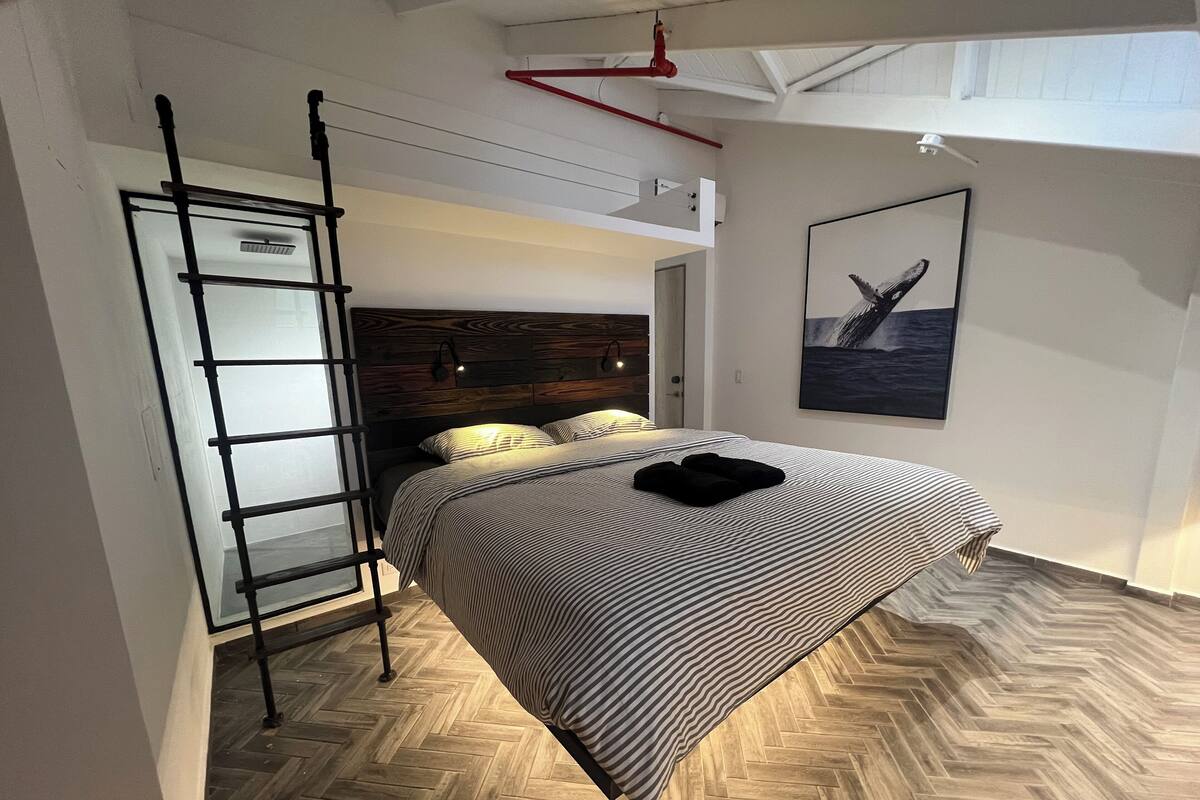 Un dormitorio con suelo de madera en espiga, una cama con ropa de cama a rayas, una toalla negra, una escalera y un cuadro enmarcado en la pared.