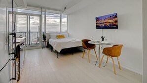 A modern hotel room with a large bed, a dining area with two chairs, a TV on the wall, and a balcony with a city view.