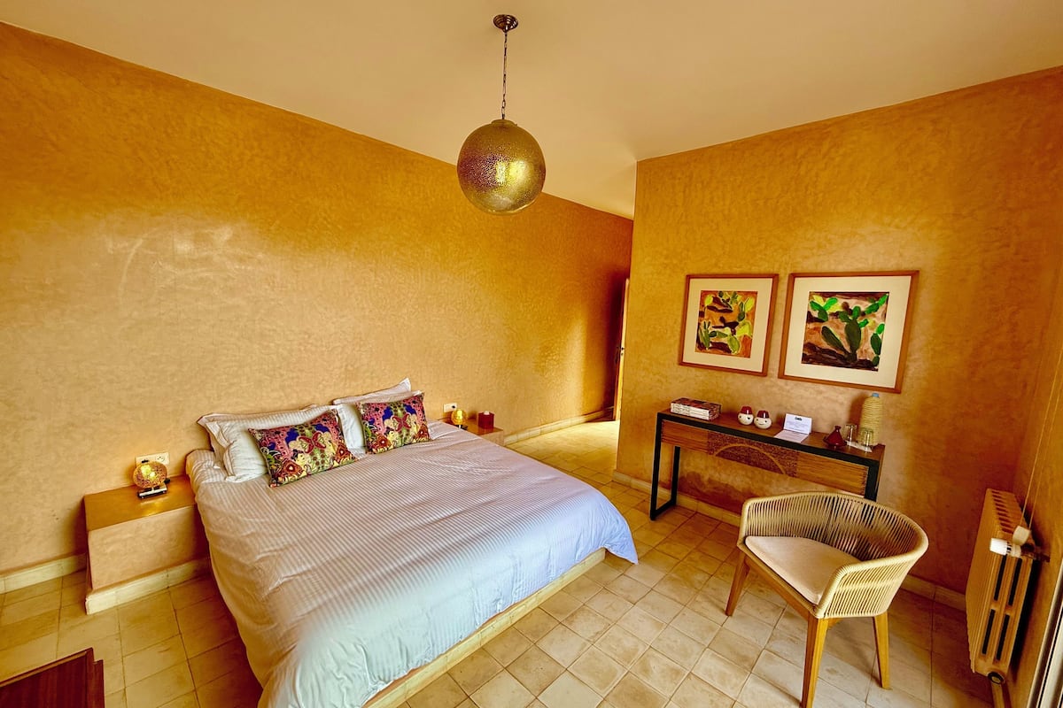 Un dormitorio con cama, mesitas de noche, escritorio, una silla y dos cuadros colgados en la pared.