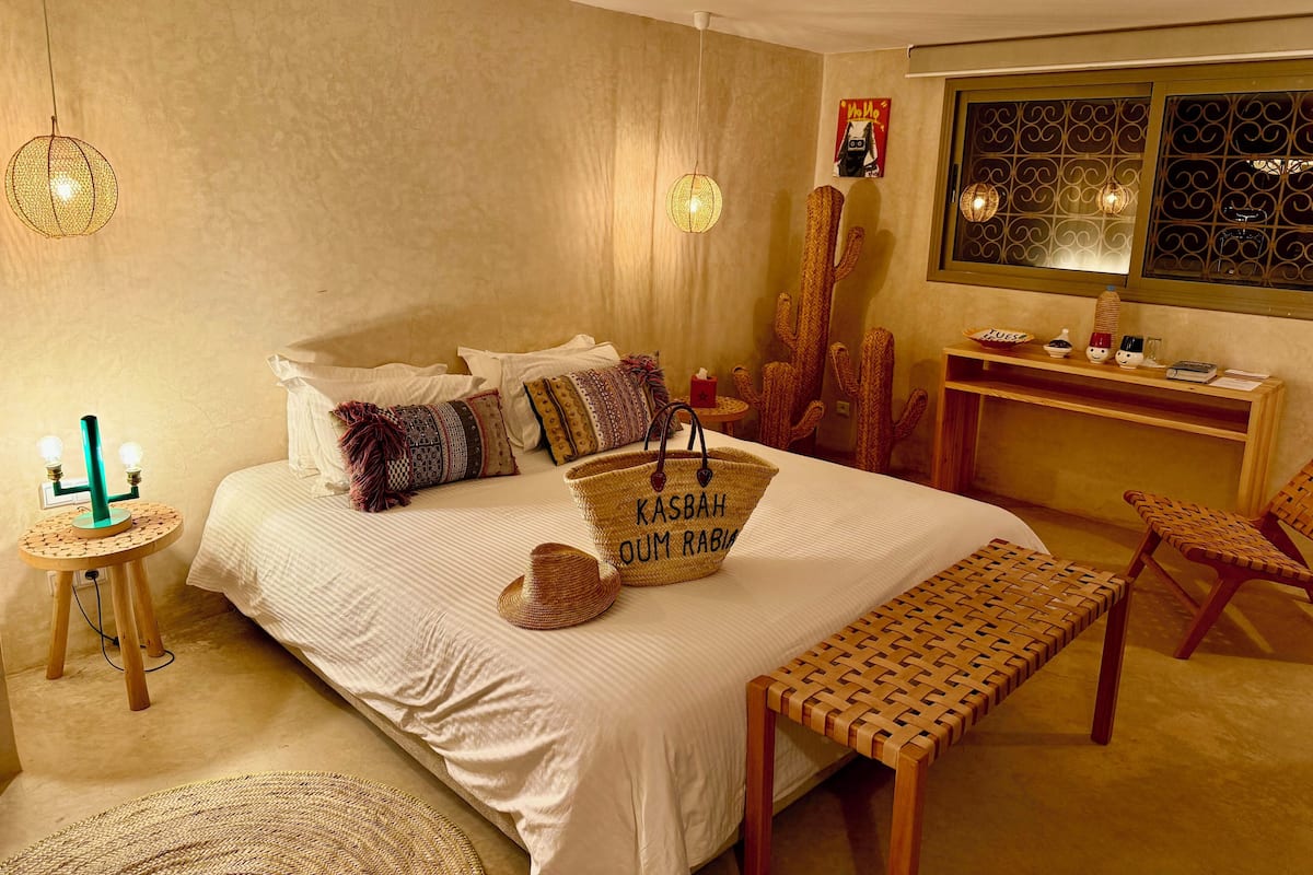 Un dormitorio con cama, un banco tejido, un cactus, un escritorio con computadora y una lámpara colgante.