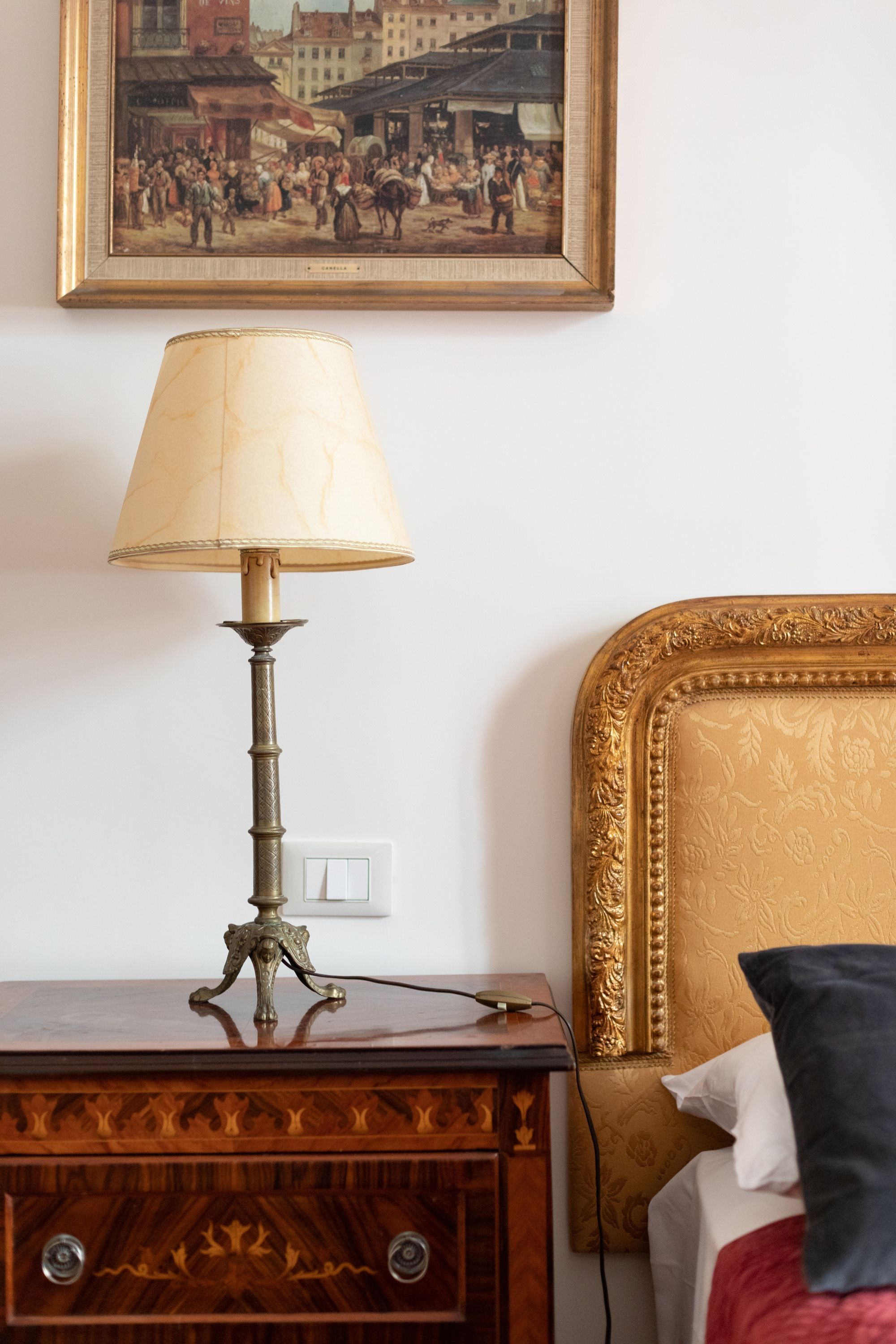 Et trænatbord med en lampe, en seng med en forgyldt sengegavl og et indrammet billede på væggen.