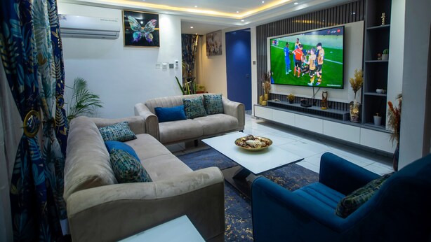 A modern living room with a large flat-screen TV, a beige sofa, a blue armchair, and a coffee table. - Fully Furnished Two Bedrooms, Modern Kitchen, Wifi (Kumasi)