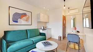 Een ruimtelijk ingerichte leefruimte met een blauwgroene bank, een klein rond tafeltje, een kitchenette en een badkamer met een spiegel.