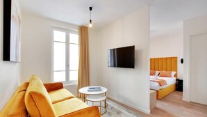 Een moderne hotelkamer met een bank, een klein rond tafeltje, een bed met oranje hoofdeinde en een flatscreen televisie die aan de muur is bevestigd.