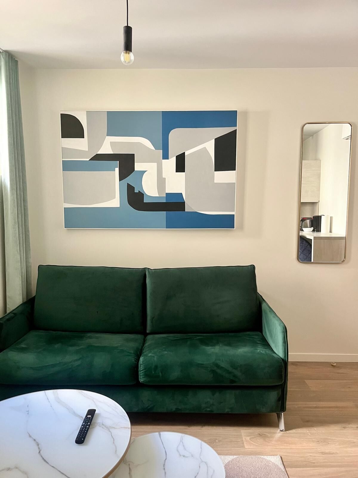 A modern living room with a green sofa, a marble coffee table, a remote control, a large abstract painting, and a mirror on the wall.