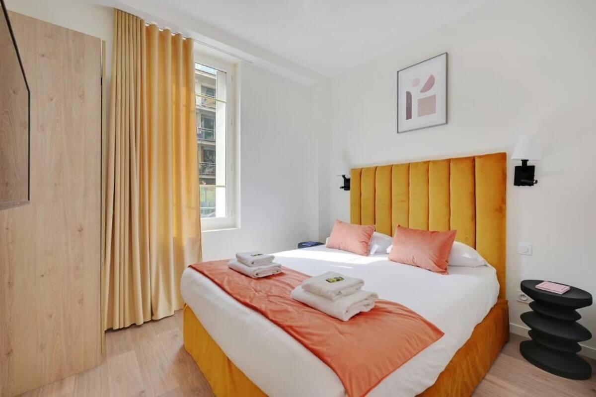 Una camera d'albergo con un letto, un comodino, una finestra con tende e un'opera d'arte appesa al muro.