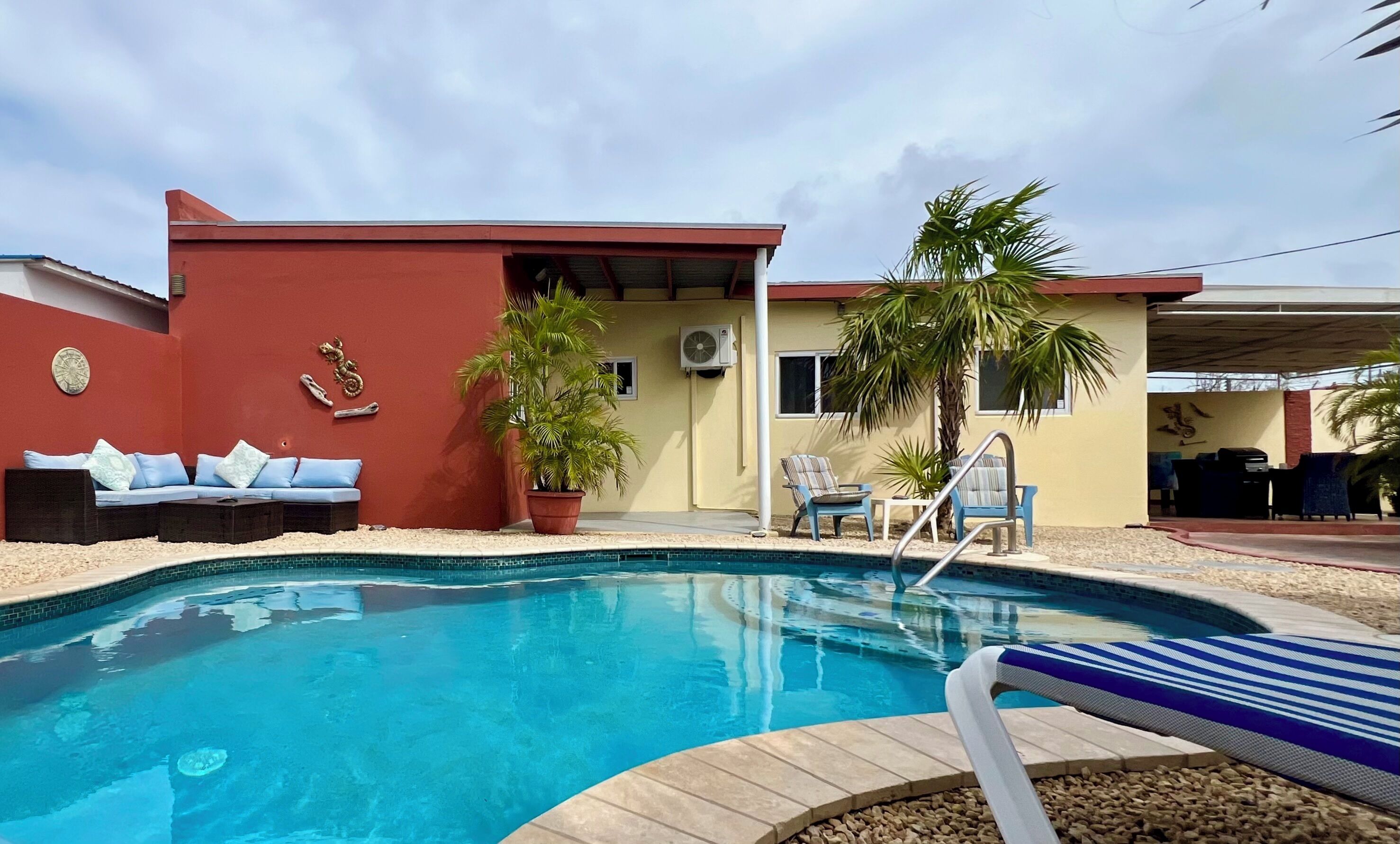 A swimming pool with lounge chairs and a small building with a red wall and a palm tree.