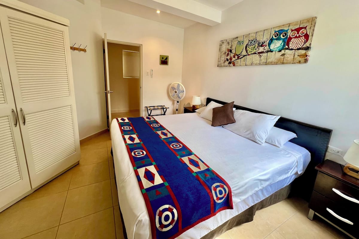 Une chambre d’hôtel comprenant un lit, une table de chevet, un ventilateur fixé au mur et une porte à persiennes.