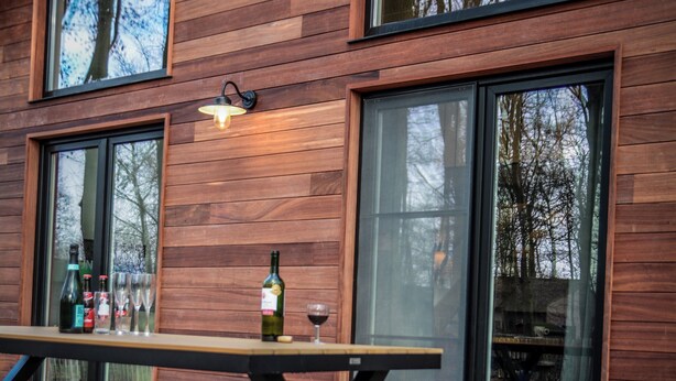 Een houten gevel met een picknicktafel en banken, grote ramen en een fles wijn op de tafel.