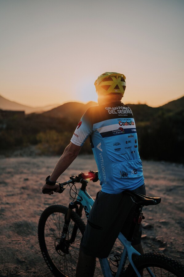 Una persona en bicicleta de montaña, con casco, parada en un sendero de tierra con un atardecer de fondo.