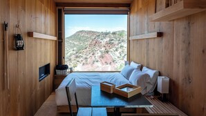 Interior de una cabaña de madera con una cama, una mesa de comedor, sillas y vistas a un paisaje montañoso.