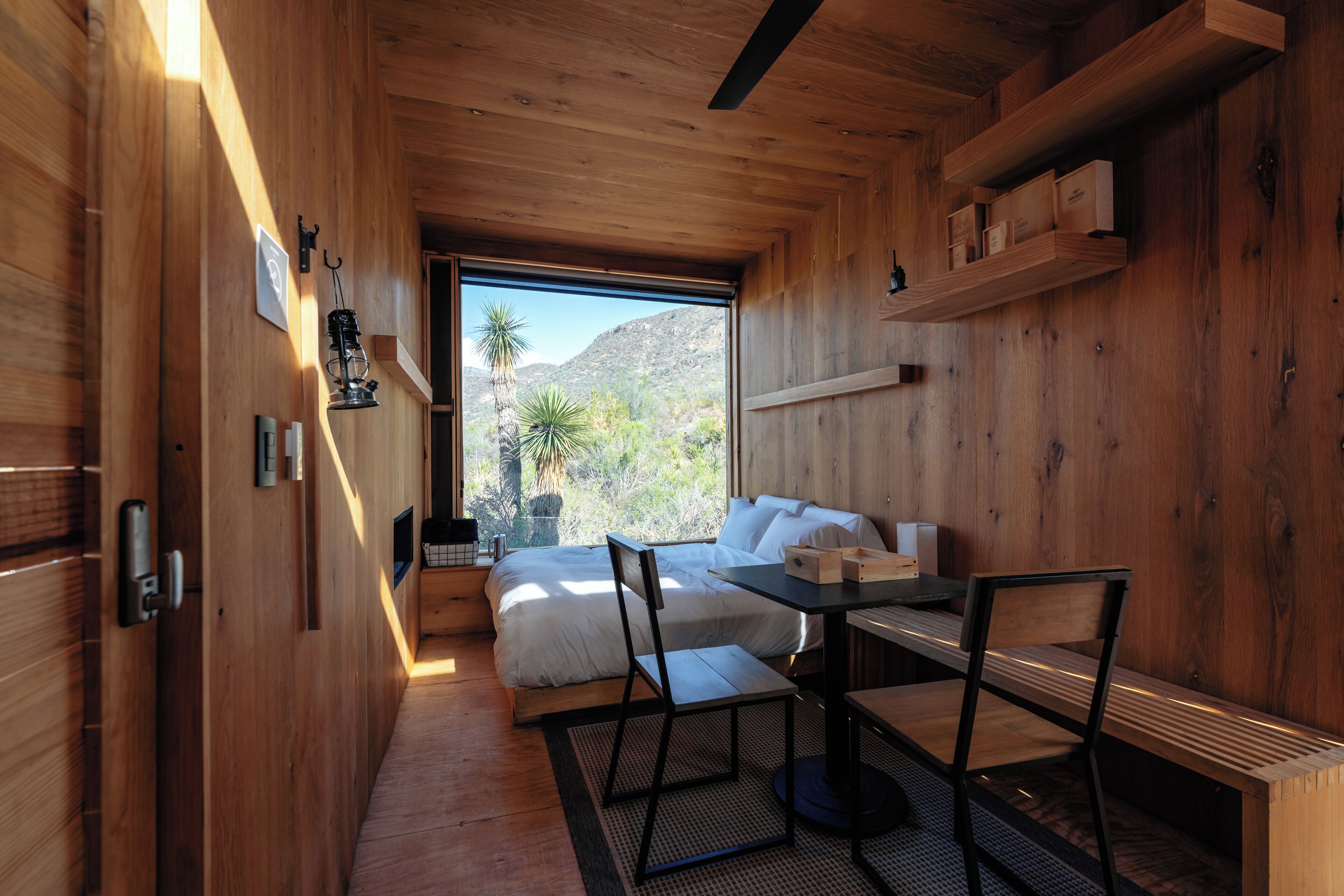 A compact room with a bed, a table, and chairs, wooden walls, a window with a view of mountains and trees, and a shelf with boxes.