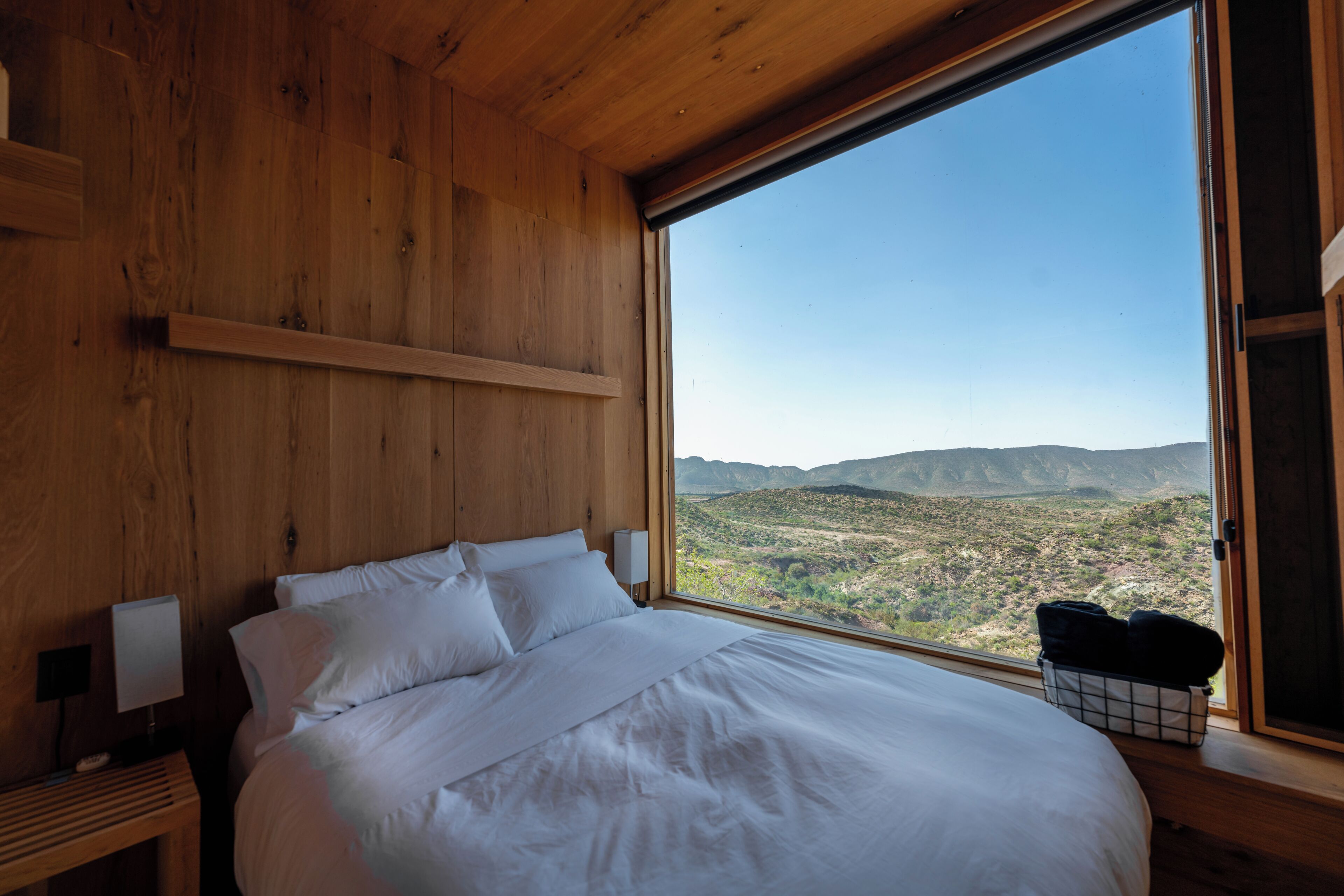 A bedroom with a large window, wooden walls, a bed with white linens, and a nightstand with a lamp.