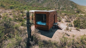 Una cabaña de madera con puerta de vidrio, rodeada de vegetación desértica y colinas.