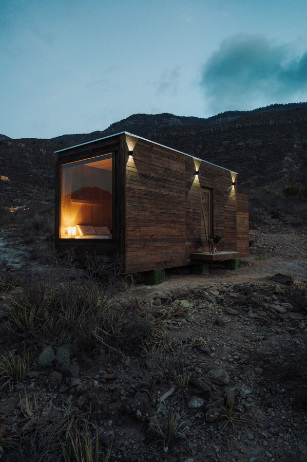 Una cabaña de madera con puerta de vidrio, una cama en el interior y un pequeño porche.