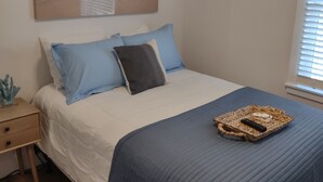 A bedroom with a bed, a nightstand, a lamp, a painting of a beach scene, and a window with blinds.