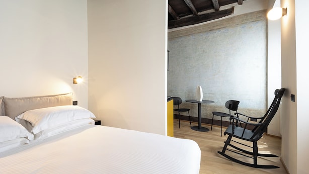 Quarto de hotel com uma cama, uma cadeira e uma mesinha.