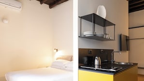 Quarto de hotel com cama, pequena cozinha equipada com pia e chaleira, e prateleira com pratos e copos.