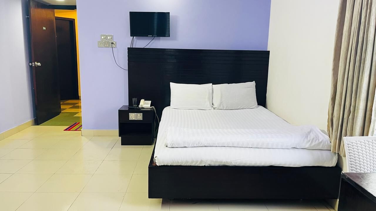 Habitación de hotel con cabecera negra, televisor de pantalla plana montado en la pared, mesita de noche con teléfono y cama con ropa de cama blanca.