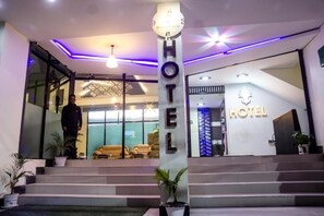 Entrada de hotel con diseño moderno, que presenta un gran letrero iluminado, una escalera que conduce al vestíbulo y una recepción con un logotipo.