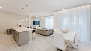 A modern kitchen with a central island, a dining area with a white table and chairs, and a living area with a sofa and a TV.