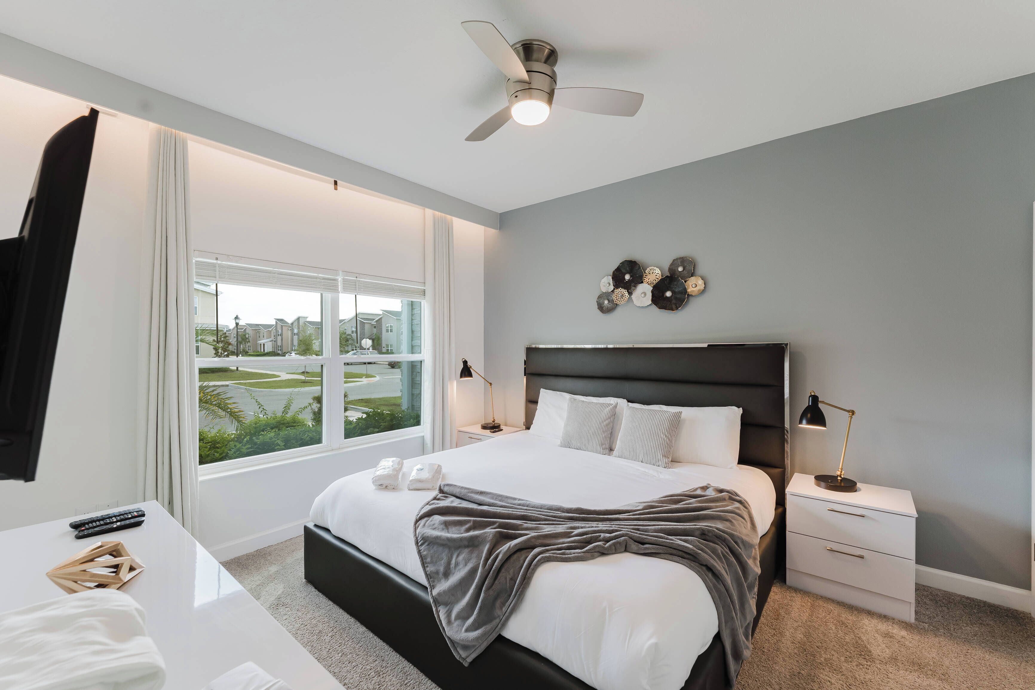 A modern bedroom with a large bed, a ceiling fan, a nightstand with a lamp, and a window with a view of a residential area.
