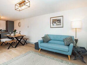 Ein modernes Wohnzimmer mit einem blauen Sofa, einem Esstisch, einer Küchenzeile und einem gerahmten Bild an der Wand.