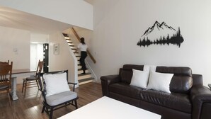 A modern living room with a brown leather sofa, a white coffee table, and a dining area with wooden chairs and a white table. A staircase with a person descending is visible in the background. - Unit 26 - Ski-in/Ski out at Mont-Sainte-Anne for 8 people (Beaupré)
