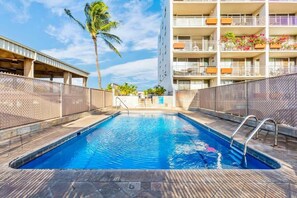 A swimming pool with a metal ladder, surrounded by a brick wall and a multi-story building with balconies.