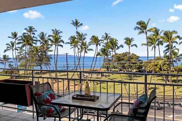 A balcony with a table and chairs, overlooking a beach with palm trees and the ocean.