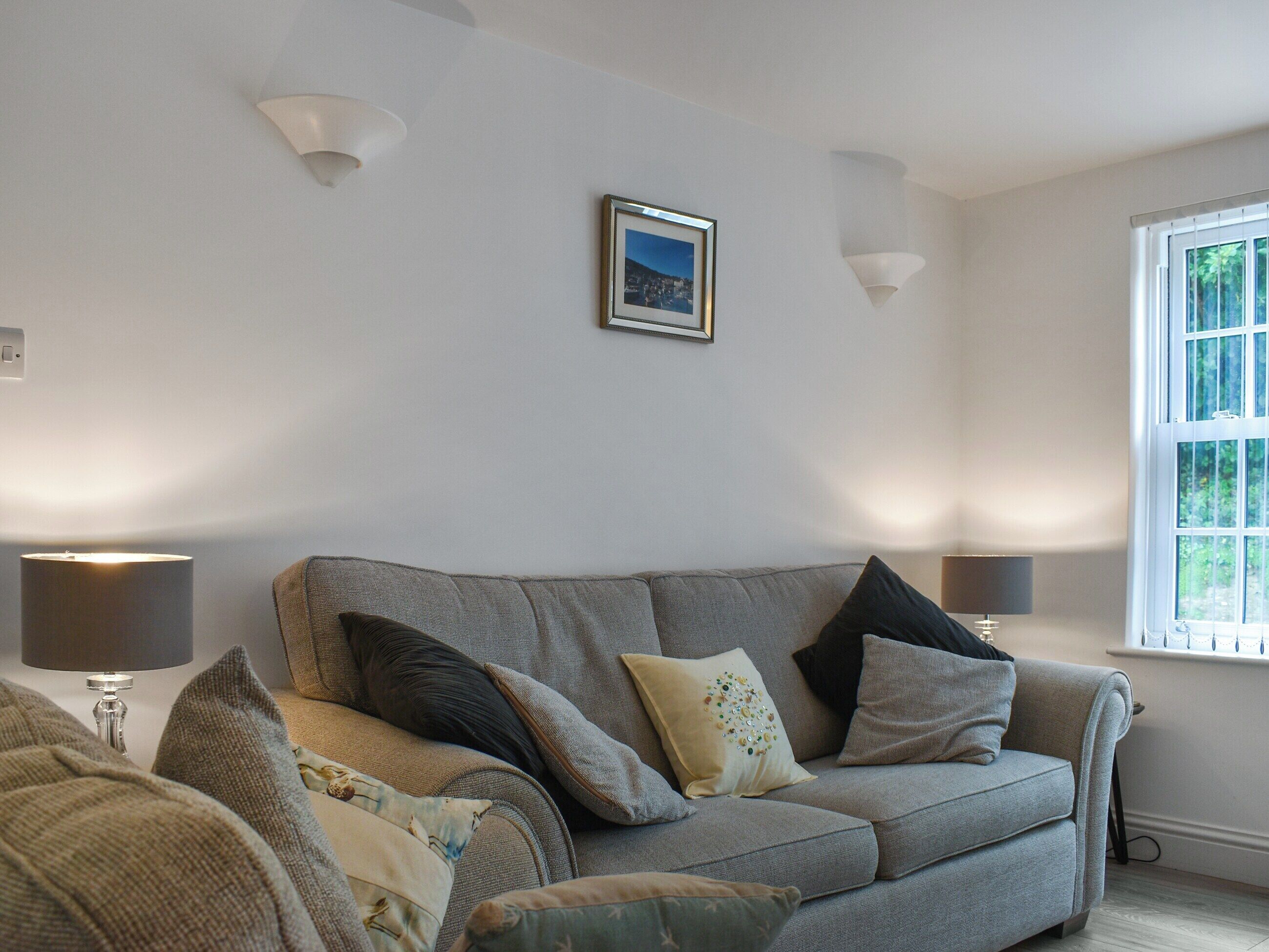 A living room with a grey sofa, multiple cushions, two wall-mounted lamps, a framed picture, and a window with blinds.
