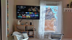 Una sala de estar con una televisión de pantalla plana montada en la pared, dos mecedoras, una estantería y una ventana con cortinas traslúcidas.