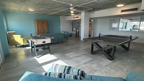 Een ruime woonkamer met een blauwe bank, een tafelvoetbaltafel en een pingpongtafel. Er is een houten kast en een raam met uitzicht.