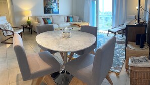 Een moderne woonkamer met een ronde marmeren tafel, grijze fauteuils, een bank met kleurrijke kussens en een schilderij aan de muur.
