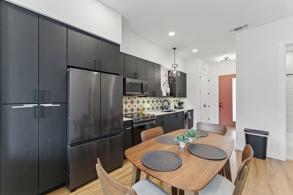 A modern kitchen with dark cabinets, a stainless steel refrigerator, and a wooden dining table with chairs. - R&R Getaway at “Freiheit Haus” with Resort Feels (New Braunfels)