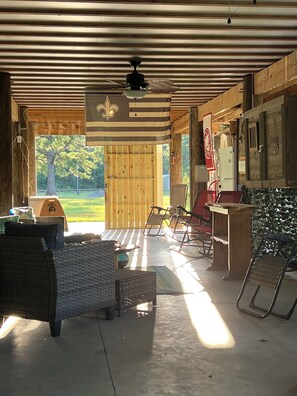 A covered outdoor area with a ceiling fan, wooden walls, a wooden door, a refrigerator, a table, and a chair.