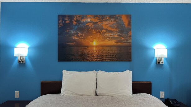 A hotel room with a bed, white pillows, a nightstand with a phone, and a sunset painting on the wall. - Extended Stay Warrenton Inn (Warrenton)