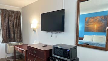 Habitación de hotel con televisor de pantalla plana montado en la pared, microondas sobre una mesita, cómoda de madera y un espejo grande que refleja una vista del atardecer.