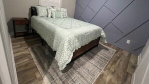 Een slaapkamer met een bed, een nachtkastje, een raam met jaloezieën, een kamerbreed tapijt met patroon en een wandpaneel met structuur.
