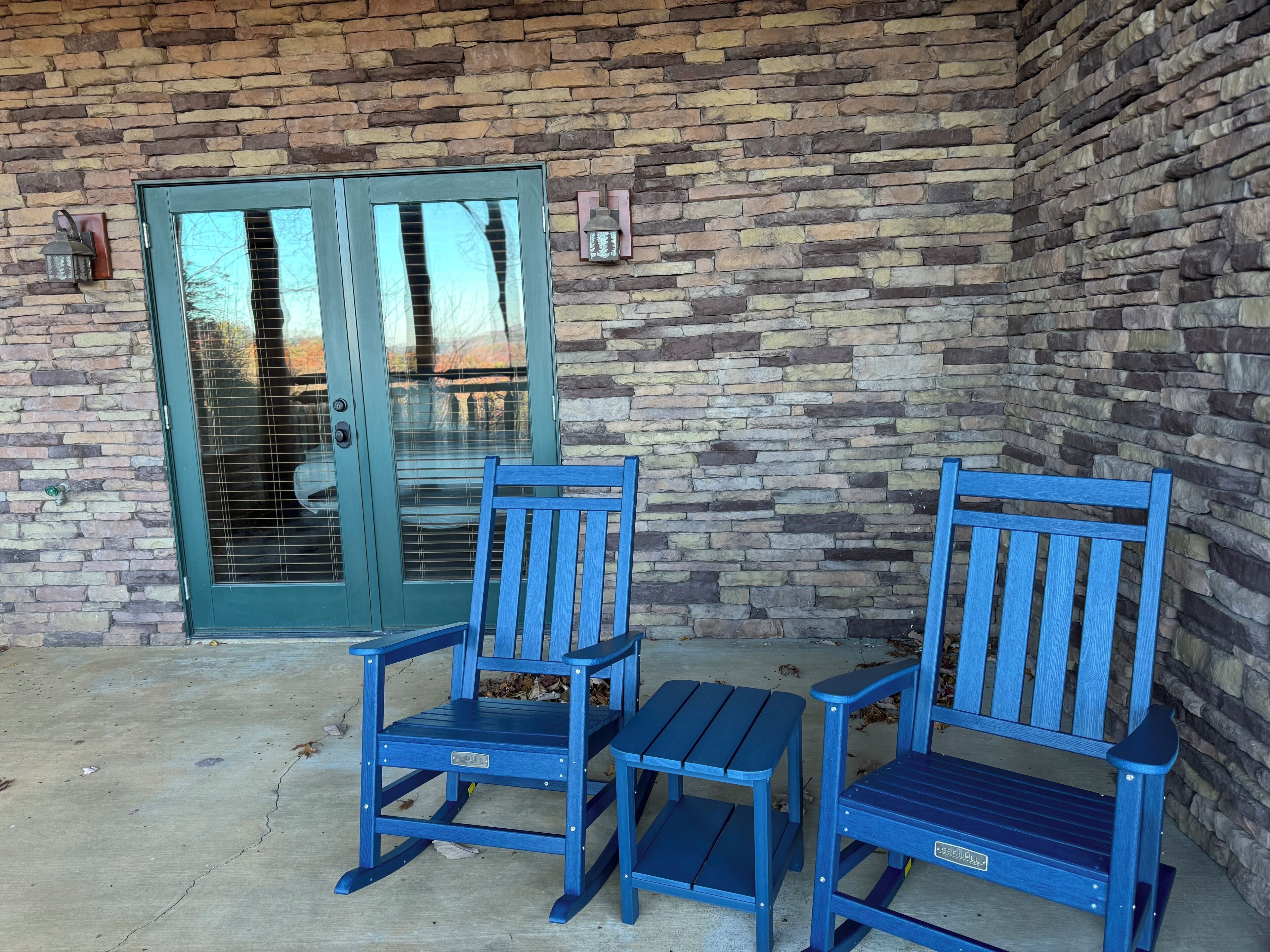 Un patio con dos mecedoras azules, una mesa, un muro de piedra y una puerta de cristal con celosía de metal.