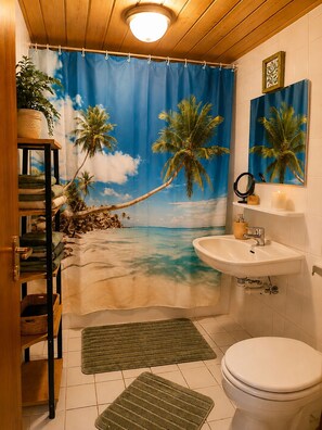 Un baño con cortina de ducha con temática playera, un lavamanos blanco, un inodoro y un estante con toallas.