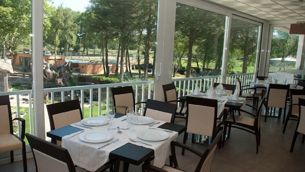 Un área de comedor al aire libre con mesas dispuestas para una comida, rodeada de sillas y con vistas a un jardín y una piscina.
