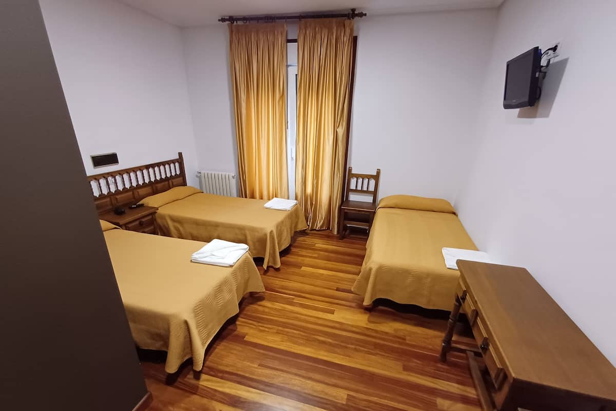 A room with four single beds, wooden flooring, a small table, a chair, and a TV mounted on the wall.