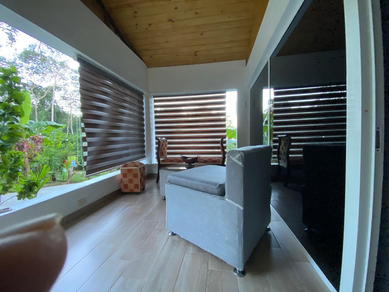Un salón moderno con un sofá gris, techo de madera y ventanas grandes con persianas.