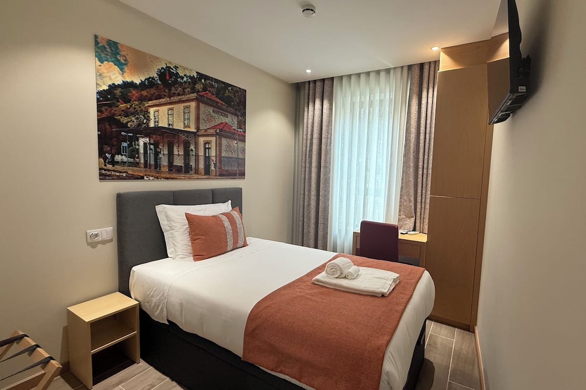 A hotel room with a bed, a nightstand, a painting, a window with curtains, and a television mounted on the wall.