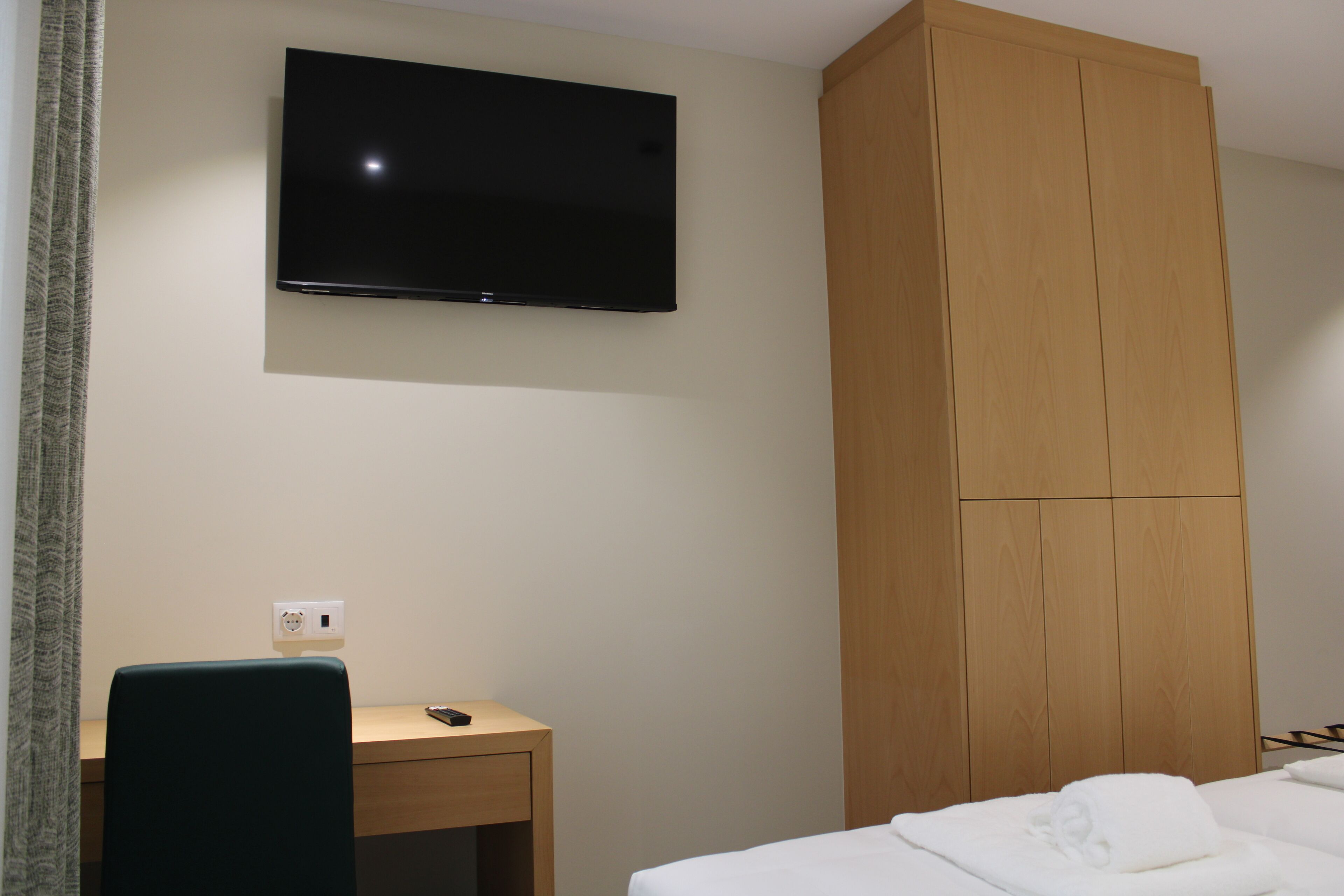 A hotel room with a bed, a desk, a television mounted on the wall, and a wooden wardrobe.