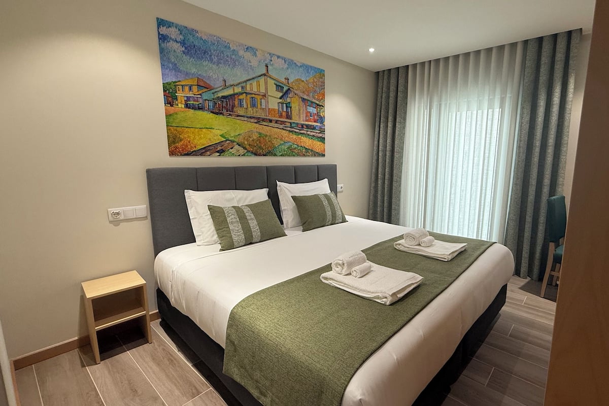 A hotel room with a large bed, a painting on the wall, and a view of the outdoors through the window.