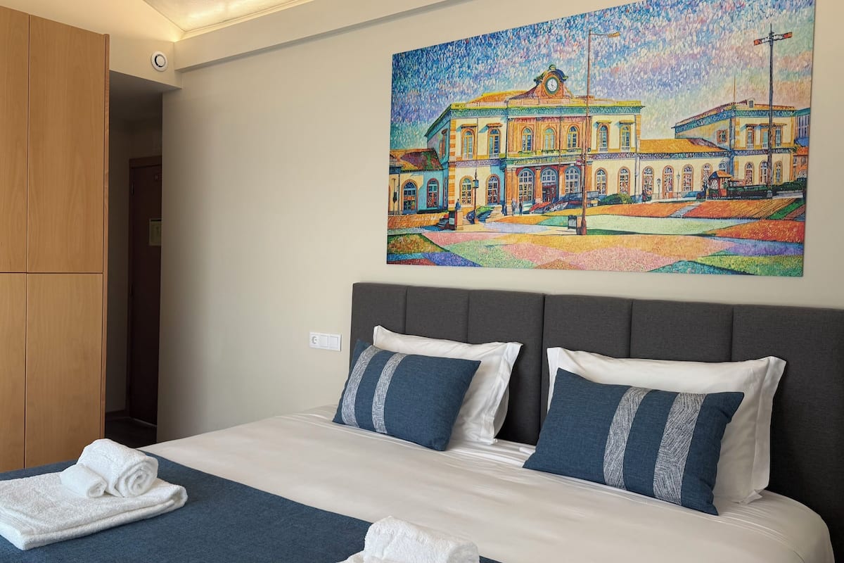 A hotel room with a large bed, two pillows, a blue blanket, and a painting of a building on the wall.